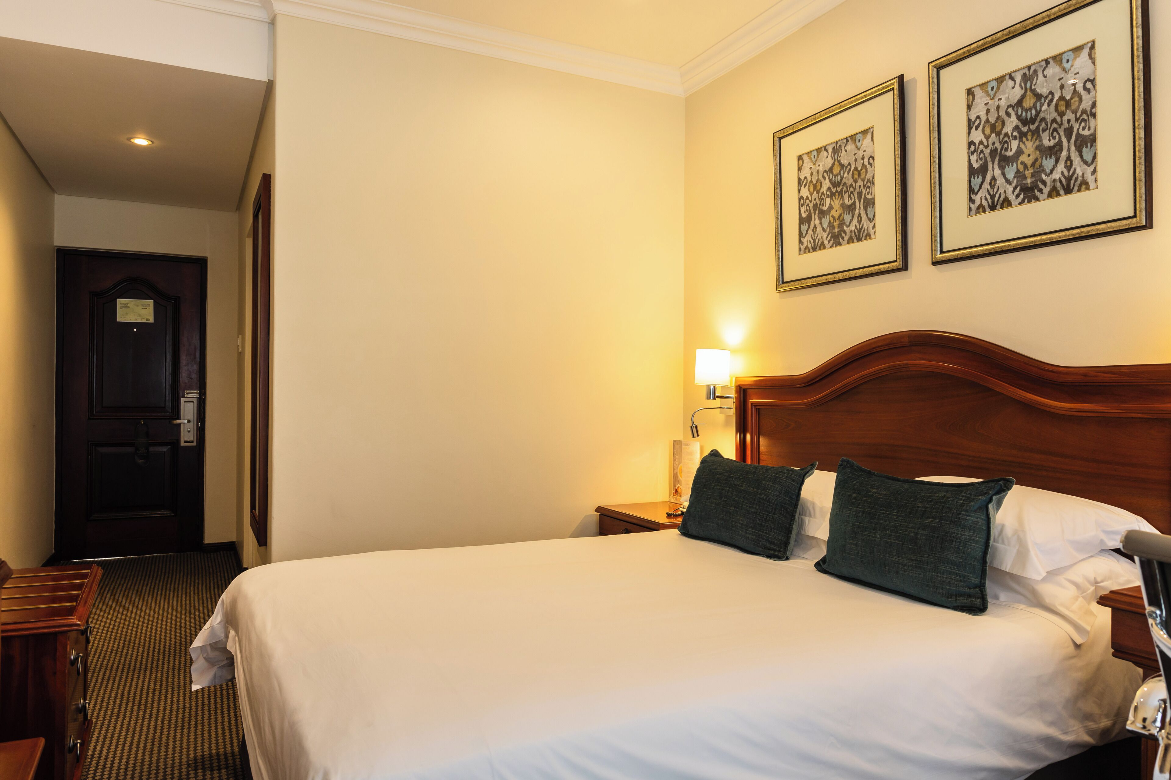 Une chambre d’hôtel avec un grand lit, une tête de lit en bois, deux tableaux encadrés au mur, une table de chevet avec une lampe et une porte sur le côté gauche.