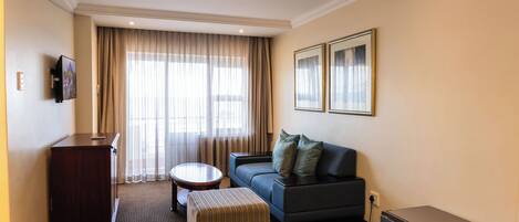 A hotel room with a sofa, ottoman, small table, TV, and a desk.
