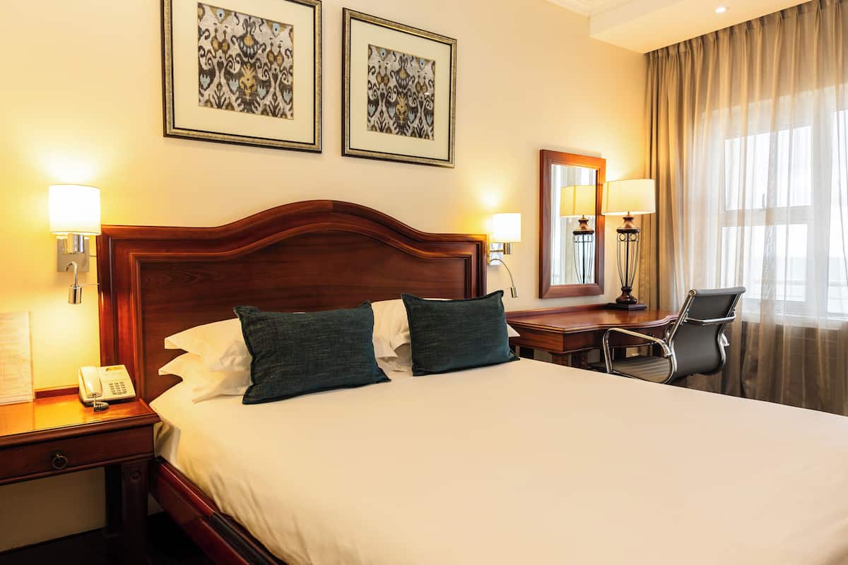 A hotel room with a large bed, two wall-mounted lamps, a desk with a chair, and two framed pictures on the wall.