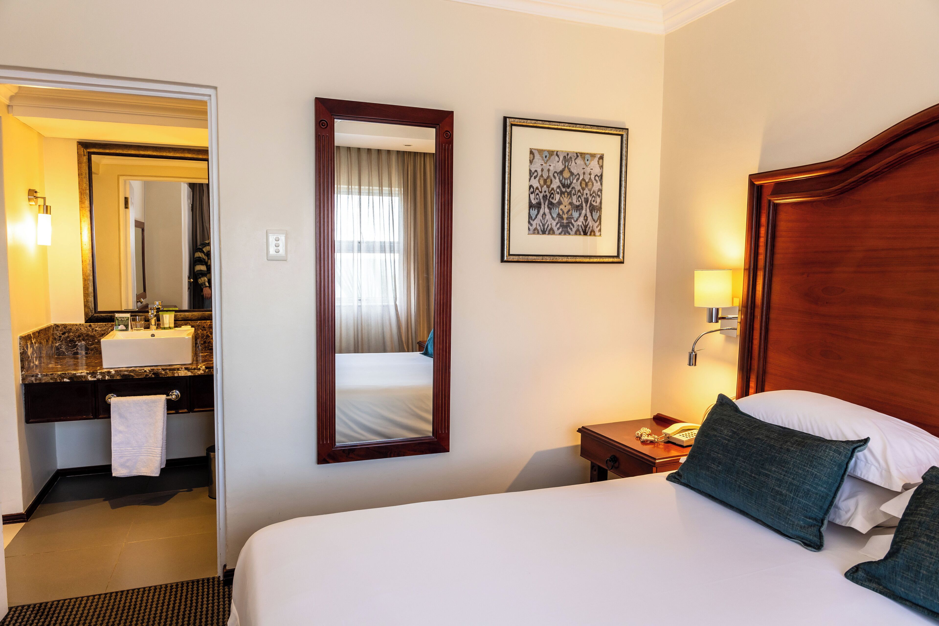 A hotel room with a large bed, a wooden headboard, a bedside table with a lamp, a framed picture on the wall, and a bathroom visible through an open door.