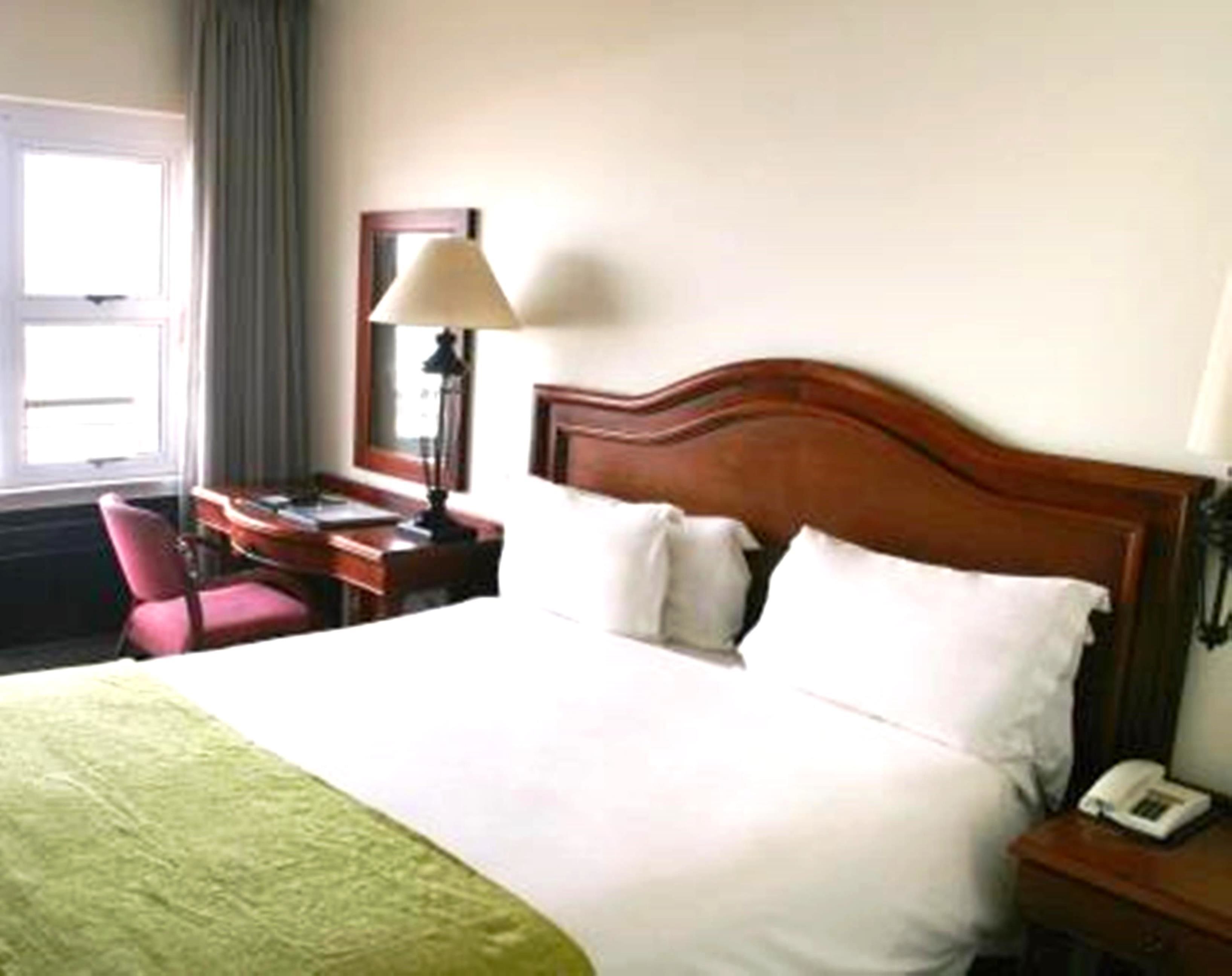 Quarto de hotel com cama de madeira, uma escrivaninha com luminária, uma cadeira e telefone no criado-mudo.