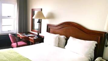 Quarto de hotel com cama de madeira, uma escrivaninha com luminária, uma cadeira e telefone no criado-mudo.