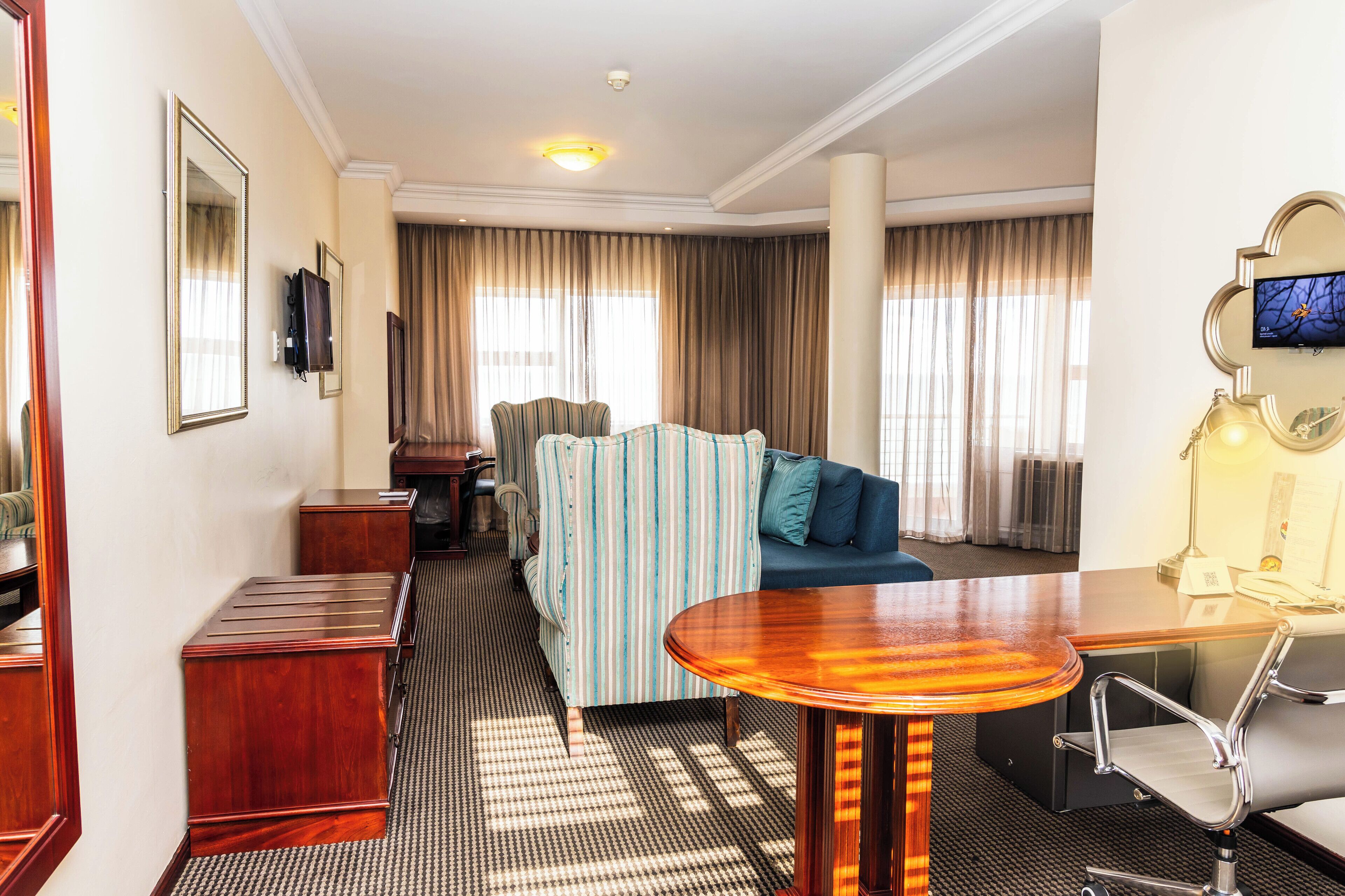 A hotel room with a striped sofa, a wooden dining table, a TV mounted on the wall, and a large window with curtains.