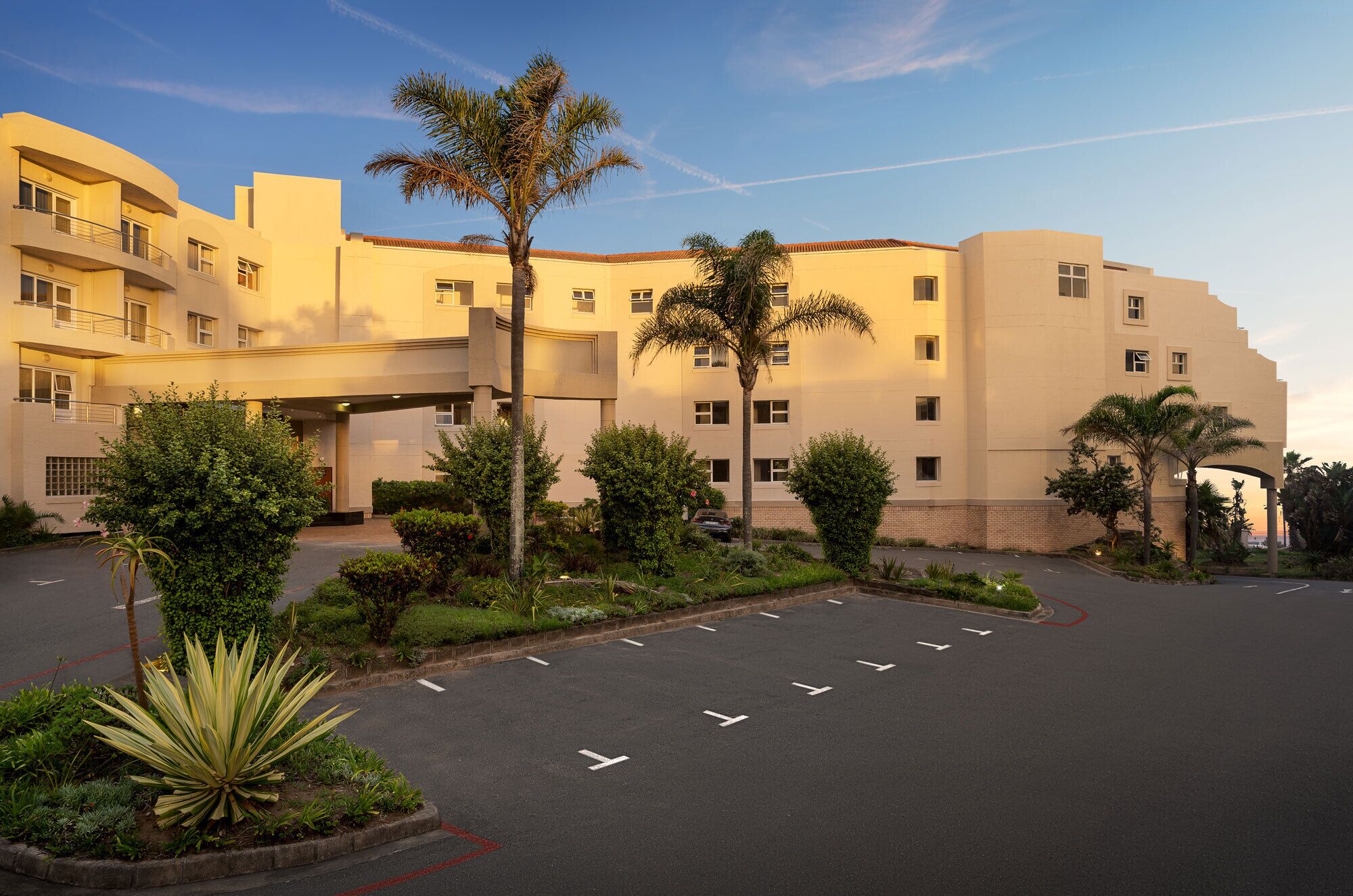 A multi-story hotel with a curved facade, a parking lot, and landscaped areas with various plants.