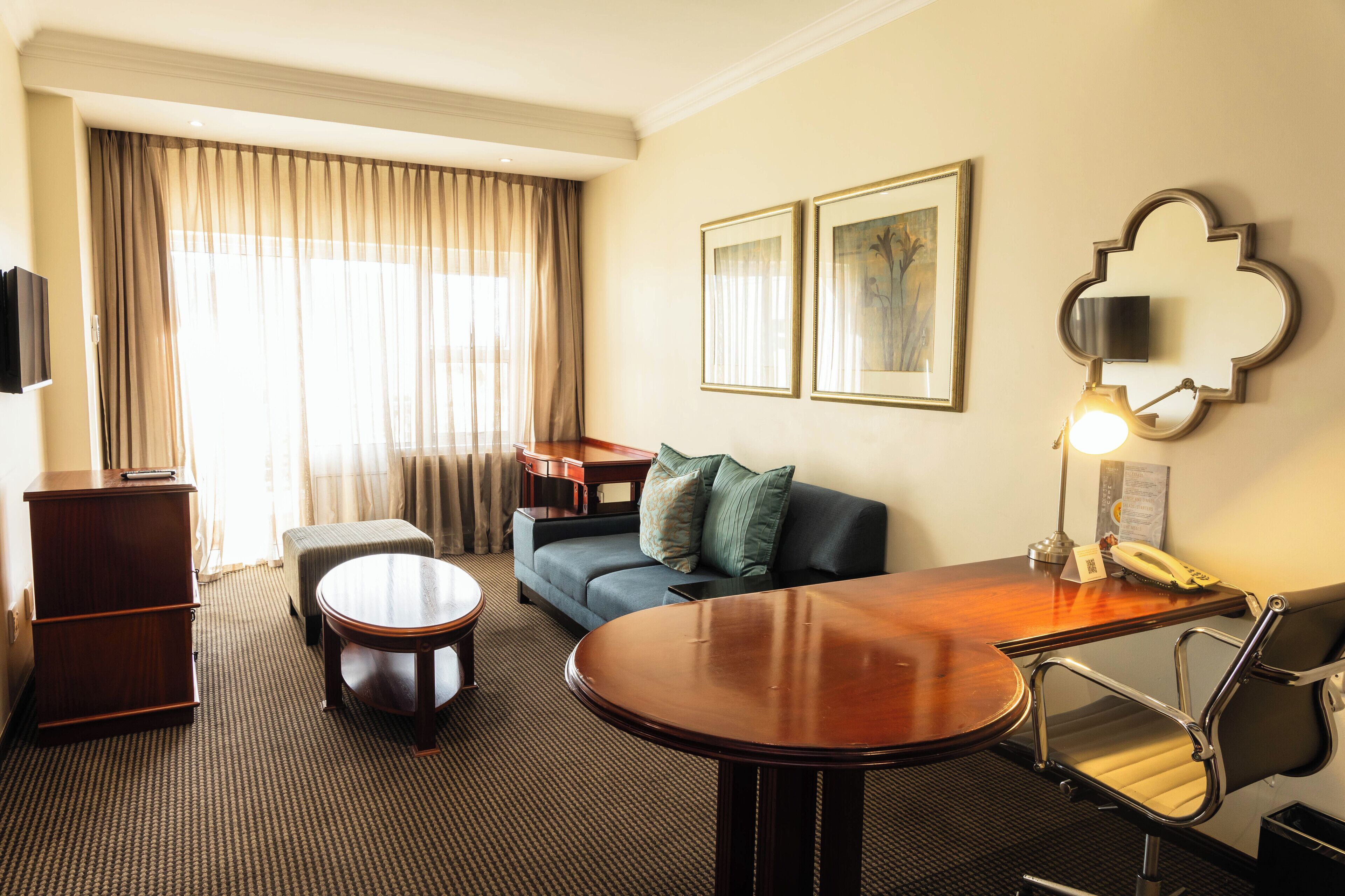 Quarto de hotel com escrivaninha de madeira, sofá, mesa redonda, cadeira, televisão e abajur.