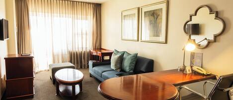 Quarto de hotel com escrivaninha de madeira, sofá, mesa redonda, cadeira, televisão e abajur.