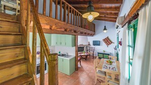 Un intérieur rustique avec un escalier en bois, une cuisine aux armoires vertes, un coin repas avec une table et des chaises, et un plafond avec des poutres apparentes et un luminaire suspendu.