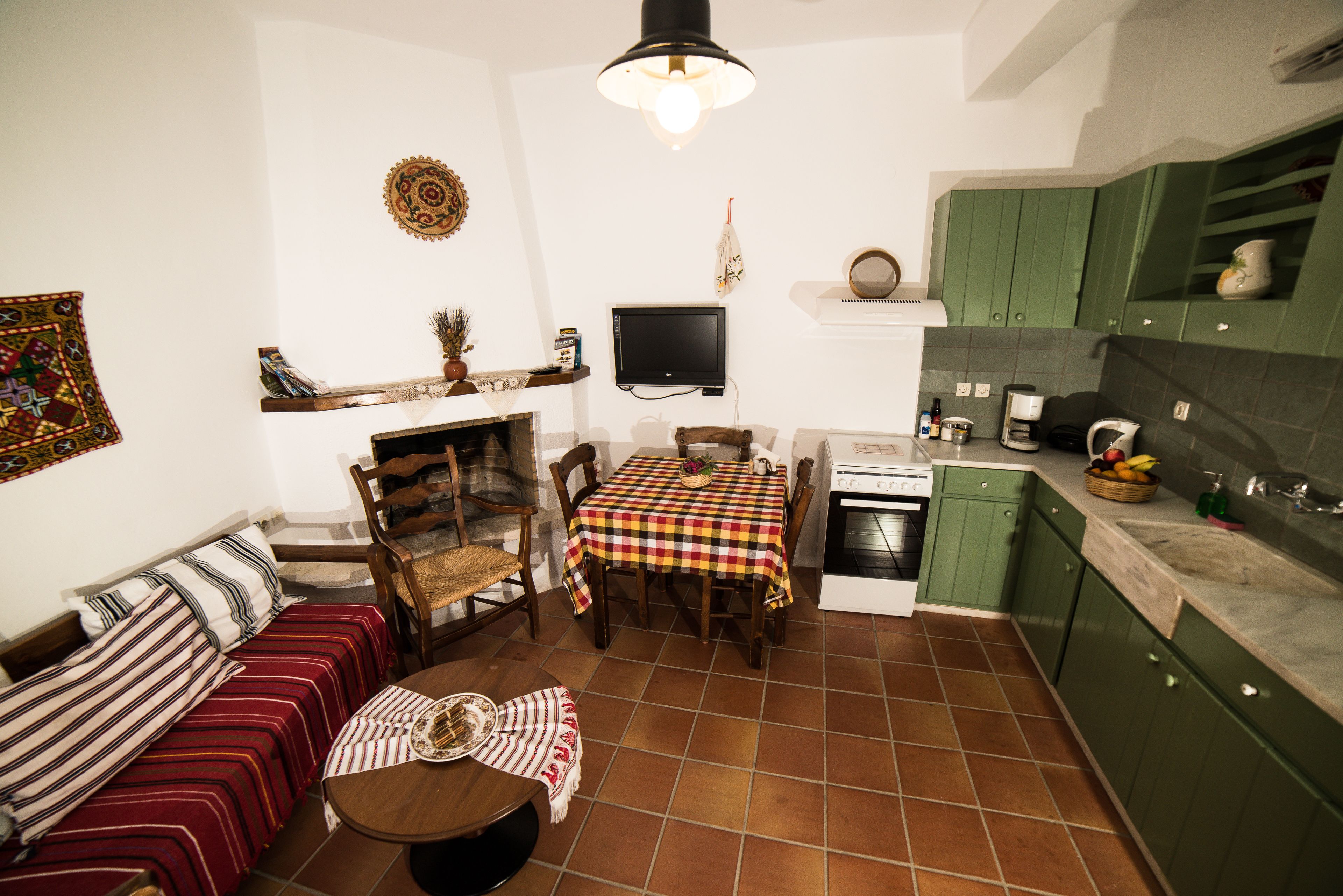 A cozy living room with a sofa, a dining table, and a fireplace. There is a kitchen area with green cabinets and a tiled floor.