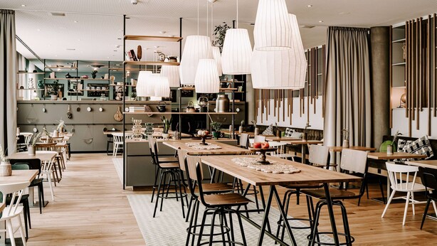 Un restaurant moderne avec des tables en bois et des chaises noires, des luminaires suspendus blancs et un espace cuisine en arrière-plan.