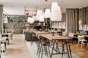 Un restaurant moderne avec des tables en bois et des chaises noires, des luminaires suspendus blancs et un espace cuisine en arrière-plan.