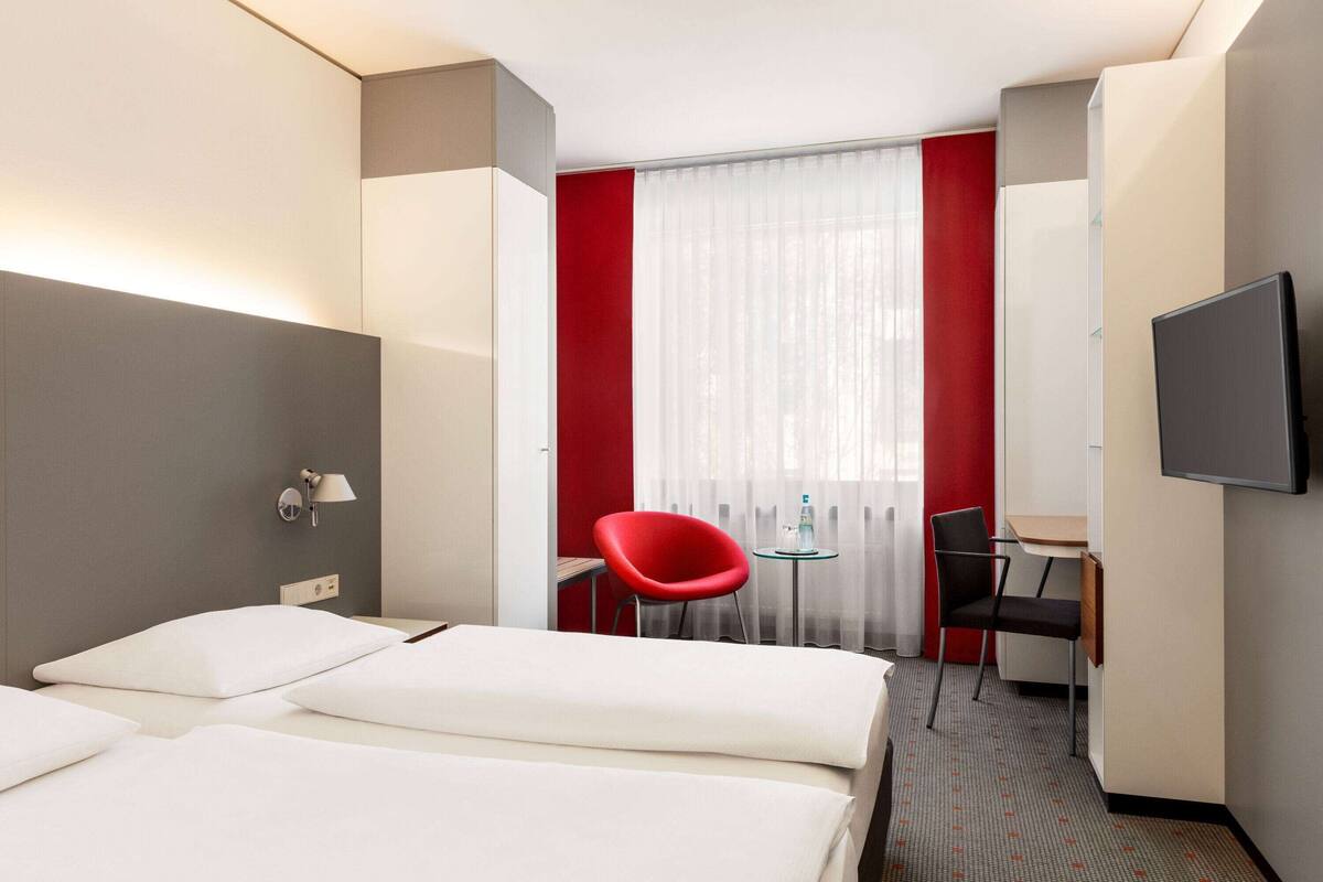 A hotel room with two beds, a desk, a TV, and a red chair.
