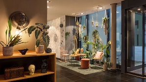 Un hall d’entrée moderne avec un comptoir d’accueil en bois, un espace détente avec des fauteuils et une petite table, ainsi que diverses plantes en pot.