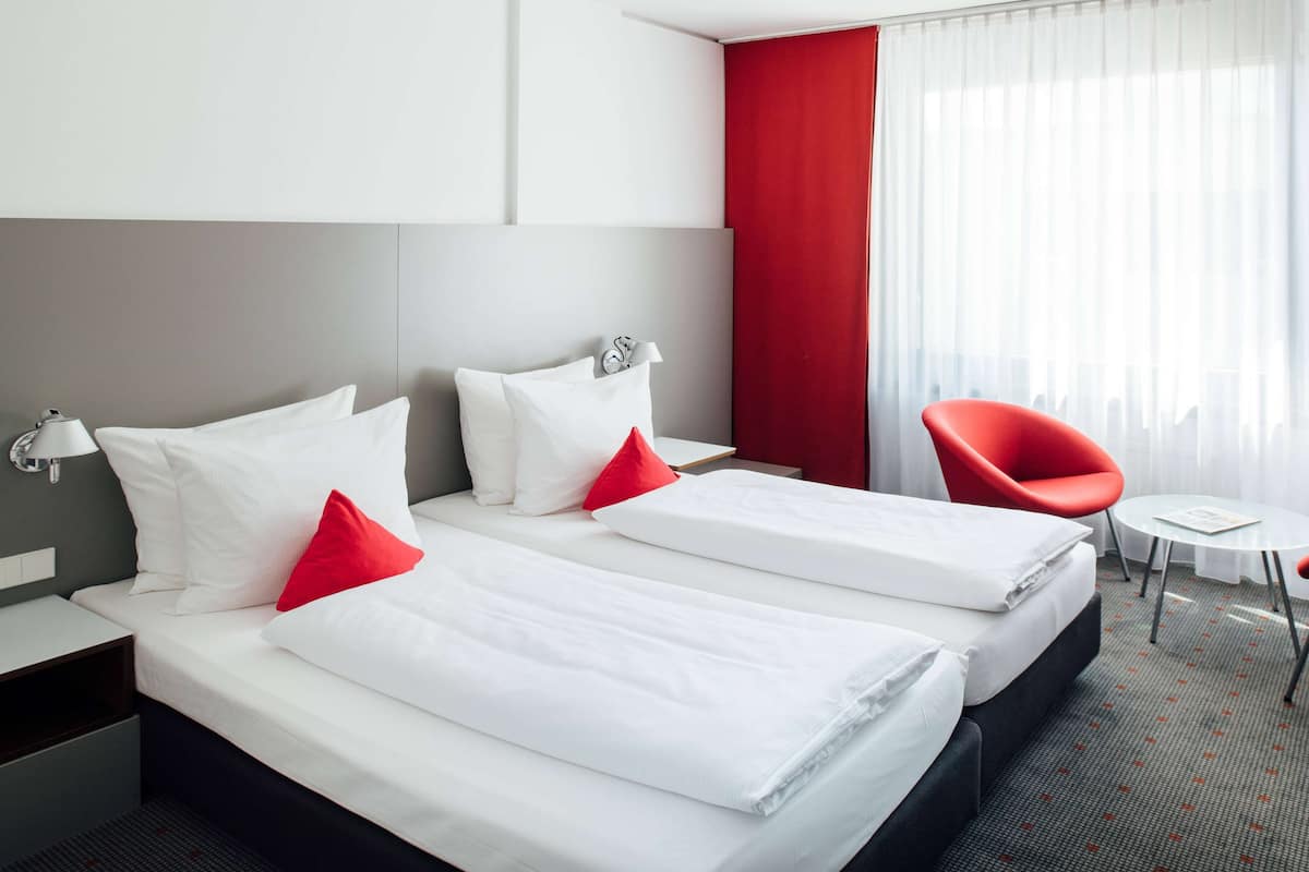 A hotel room with two beds, a red chair, a small table, and a window with curtains.
