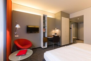 A hotel room with a bed, a red chair, a desk, a television, and a small table with a red book. - Vienna House Easy by Wyndham Stuttgart (Stuttgart)