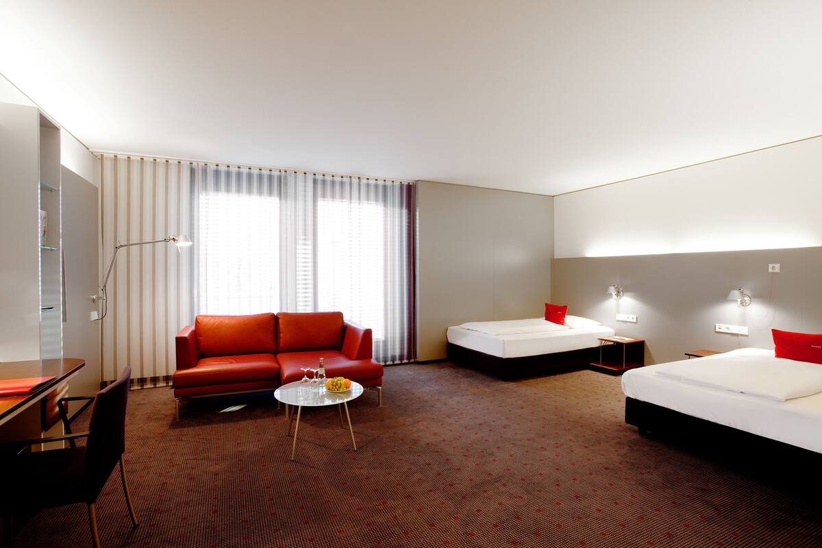 A hotel room with a red sofa, two beds, a small table with a bottle and glasses, and a lamp.