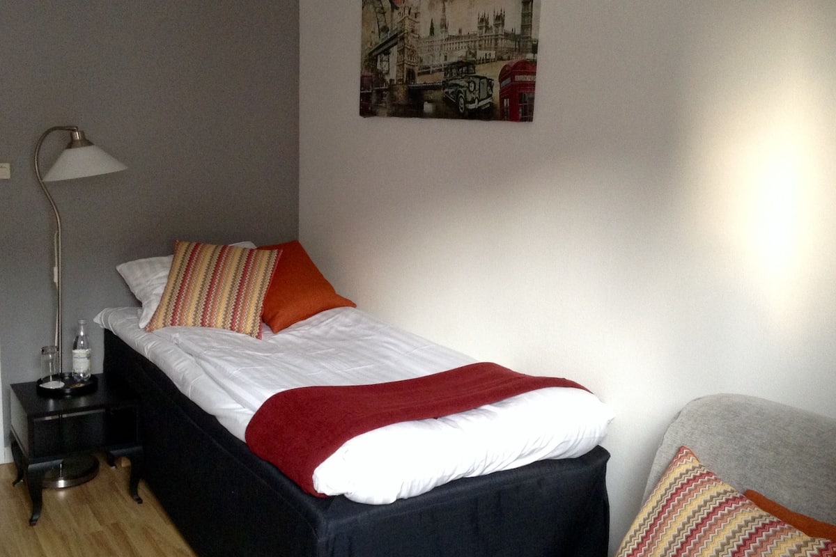 Uma cama bem arrumada com lençóis brancos e manta vermelha, luminária de cabeceira, mesa pequena com garrafa e copos, e um quadro na parede de um edifício histórico.