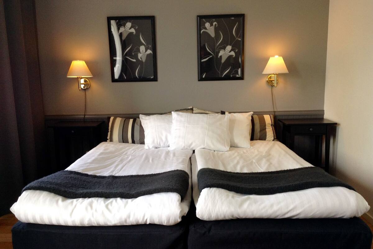 Uma cama de casal com roupa de cama preta e branca, duas luminárias fixadas na parede e duas obras de arte com motivos florais emolduradas.