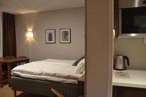 A hotel room with a bed, a desk, a chair, a wall-mounted light, and a kettle on the counter.