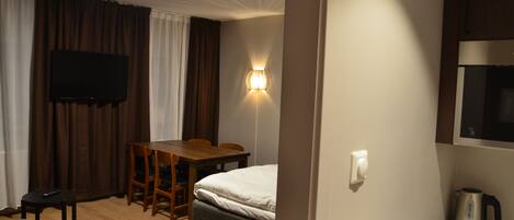 Habitación de hotel con cama, escritorio, silla, televisor, lámpara de pared, tetera y una mesita con control remoto y llaves.
