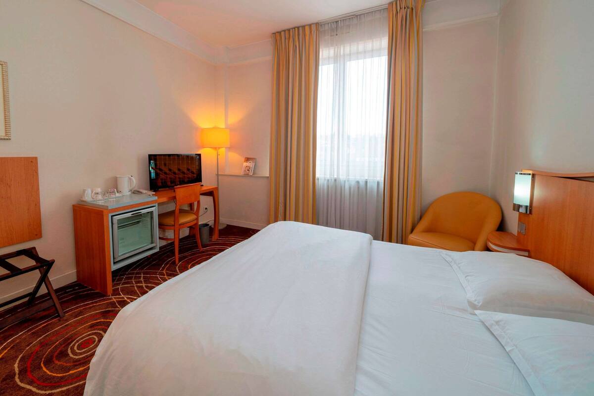 Ein Hotelzimmer mit einem großen Bett, einem Schreibtisch mit Computer, einem Sessel, einem Fernseher und einer Lampe.