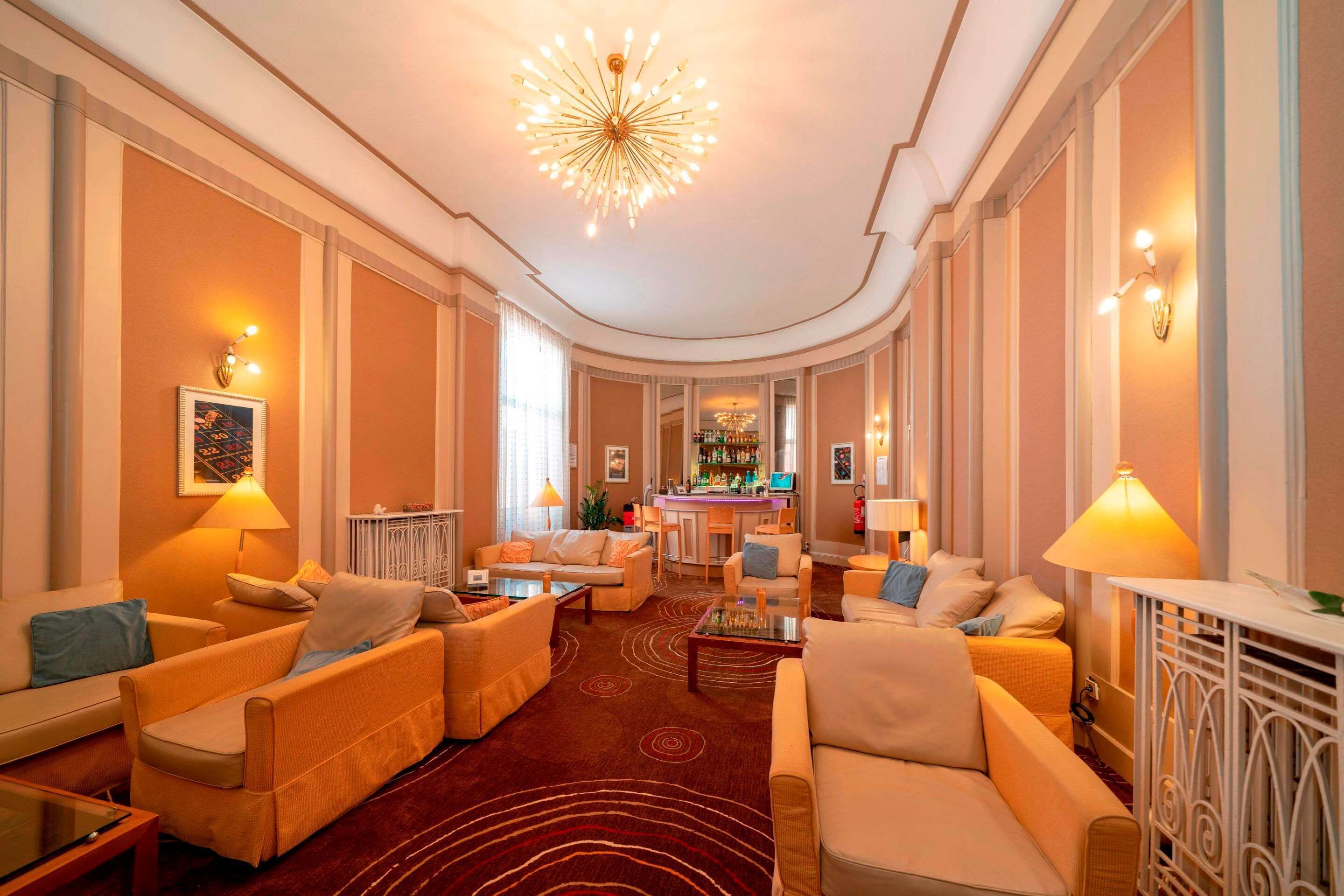 Een ruime hotellobby met een bar in het midden, diverse zithoekjes en een decoratieve plafondlamp.