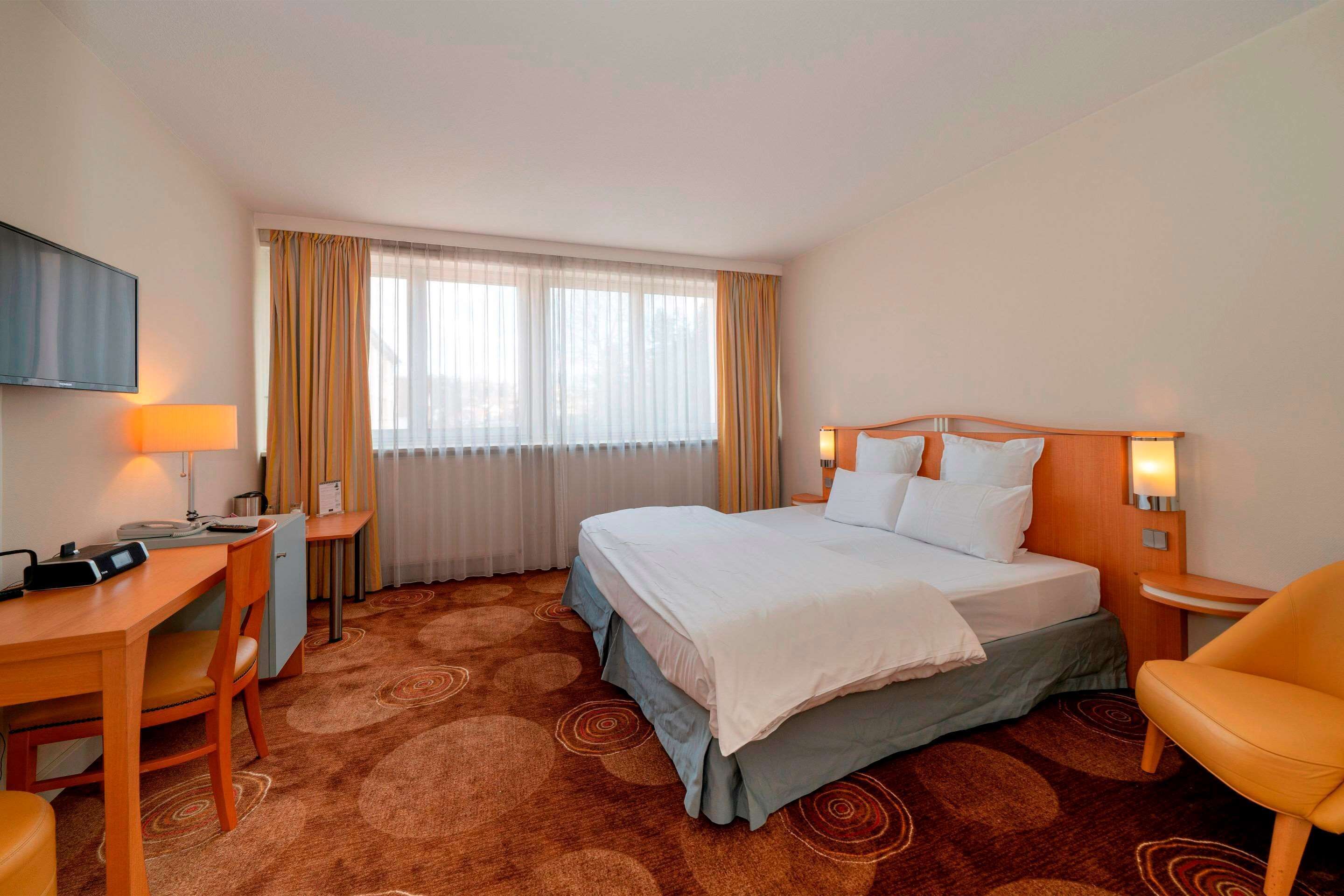 A hotel room with a bed, a desk, a chair, a TV, and a window with curtains.