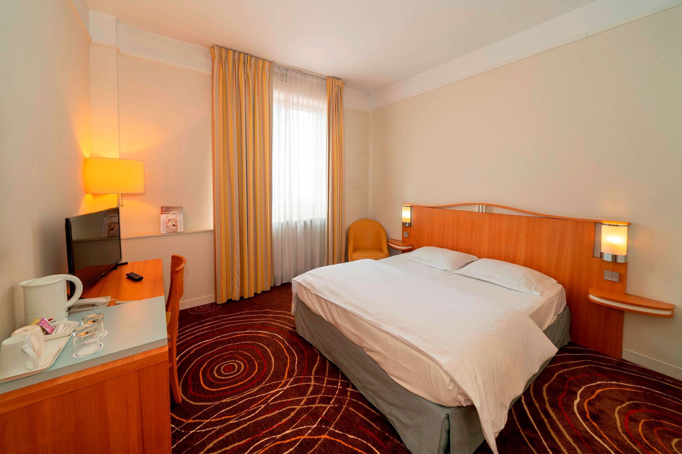 Ein Hotelzimmer mit Bett, Schreibtisch mit Lampe, Stuhl, Fernseher und Fenster mit Vorhängen.