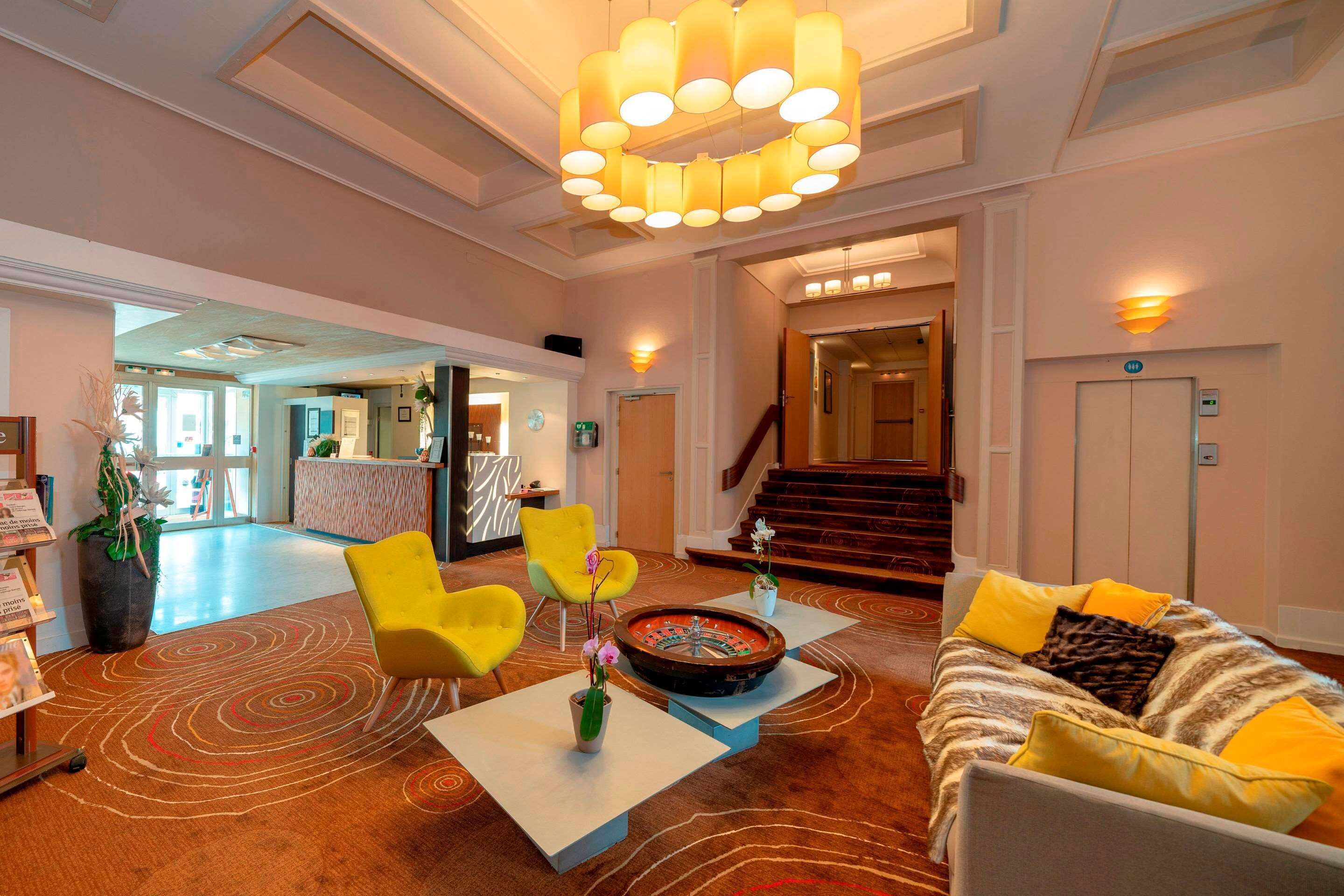 A lobby with a round table, yellow chairs, a sofa with yellow cushions, a large chandelier, and a staircase in the background.