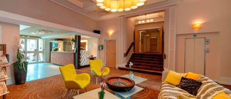 A lobby with a round table, yellow chairs, a sofa with yellow cushions, a large chandelier, and a staircase in the background.