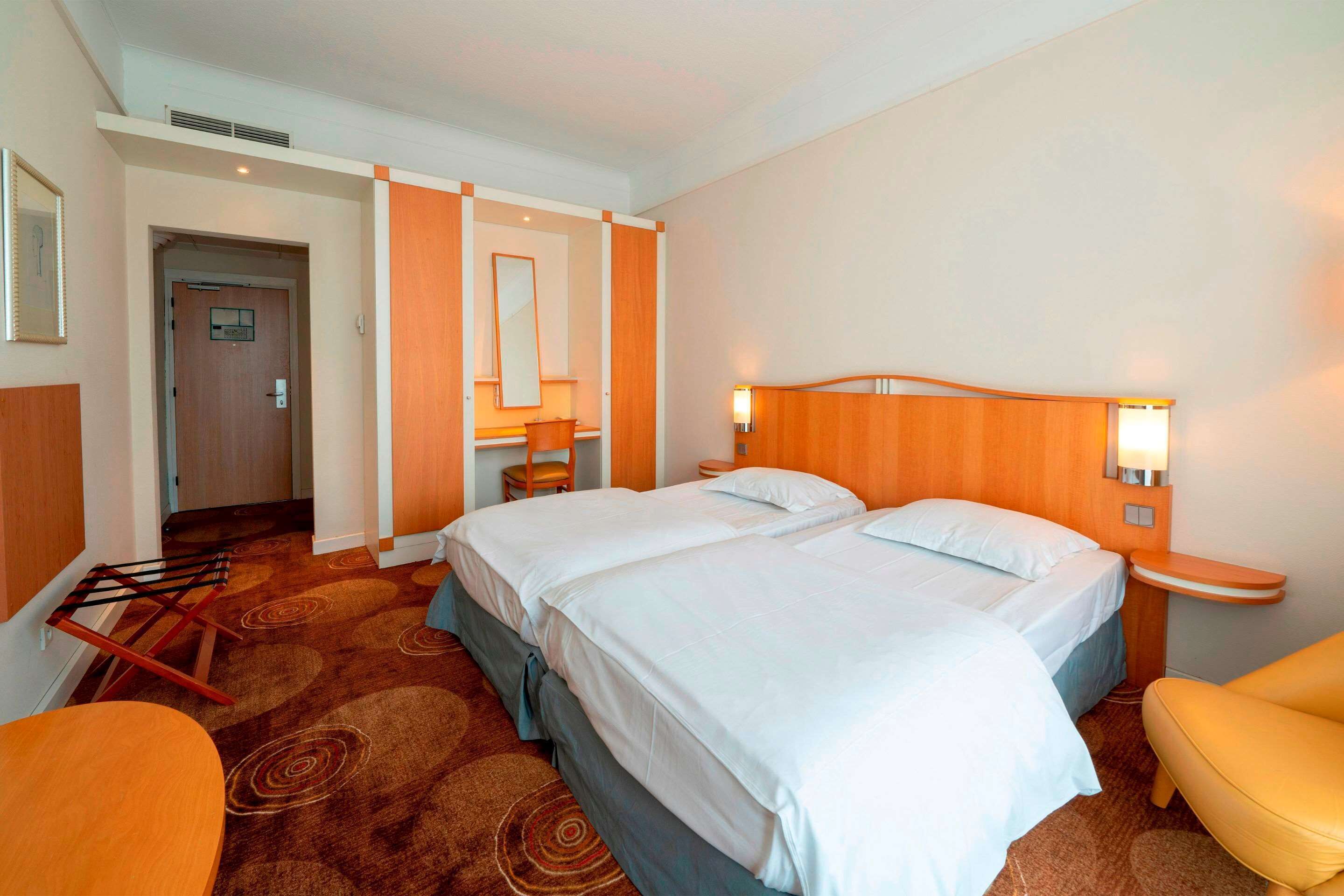 A hotel room with two beds, a sitting area with a chair, a small table, and a bathroom with a mirror and sink.