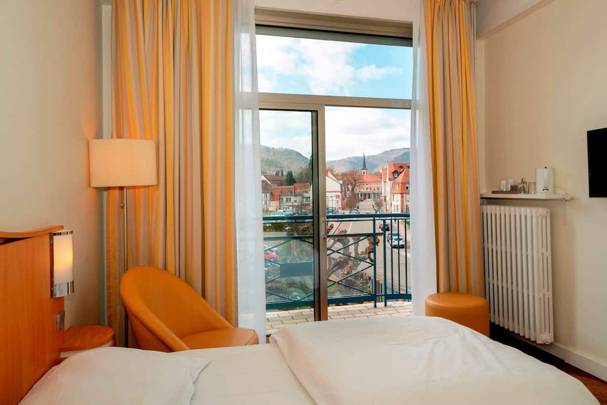 Ein Hotelzimmer mit Bett, Stuhl, Lampe, Blick auf die Stadt durch ein großes Fenster und einer Heizung.