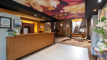 A hotel reception area with a wooden reception desk, a large floral mural on the ceiling, a staircase, and a seating area with a sofa and chairs.
