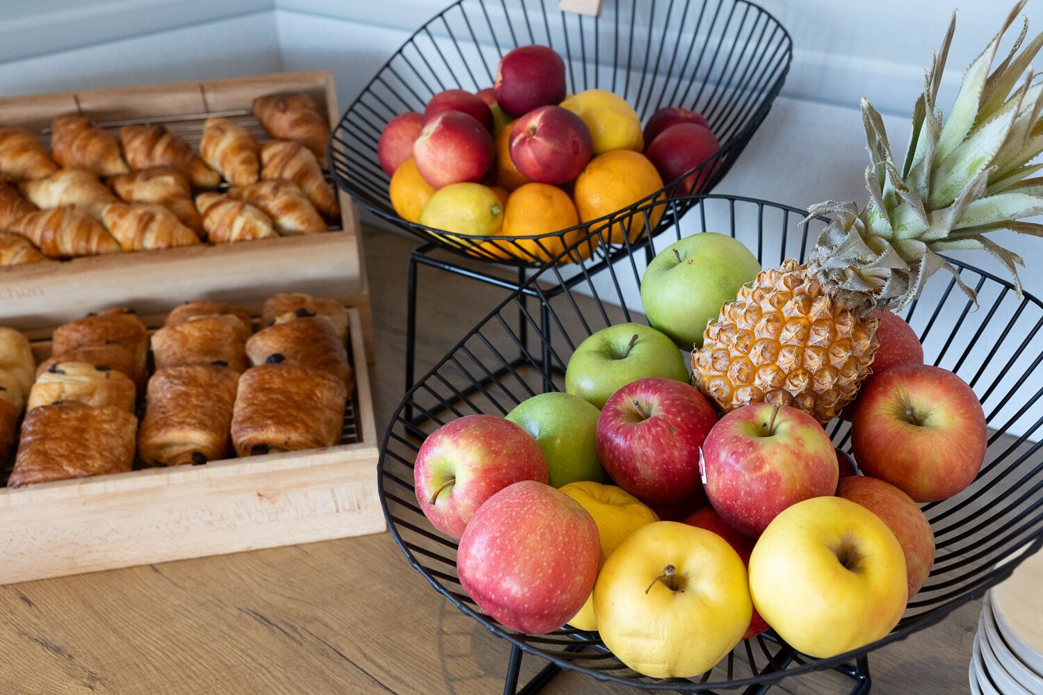 A kitchen counter with a fruit bowl containing apples, oranges, and a pineapple, and a wooden box with pastries.
