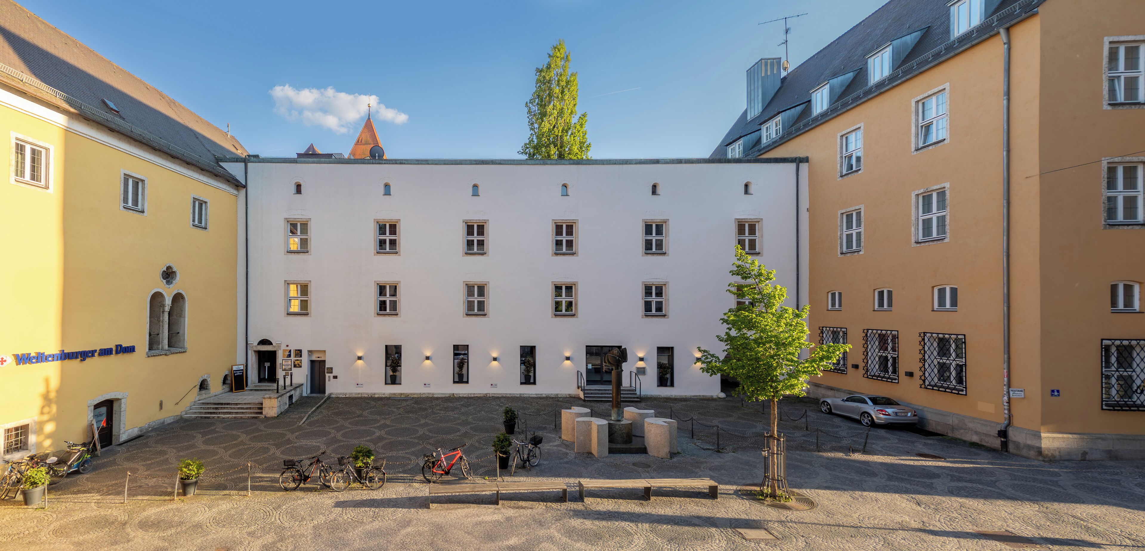 A courtyard with a white building, bicycles, and a car parked nearby.