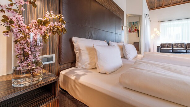 A hotel room with a large bed, a wooden headboard, a vase with flowers, and a sofa in the background. - Hotel Regensburg Herzogshof am Dom (Regensburg)