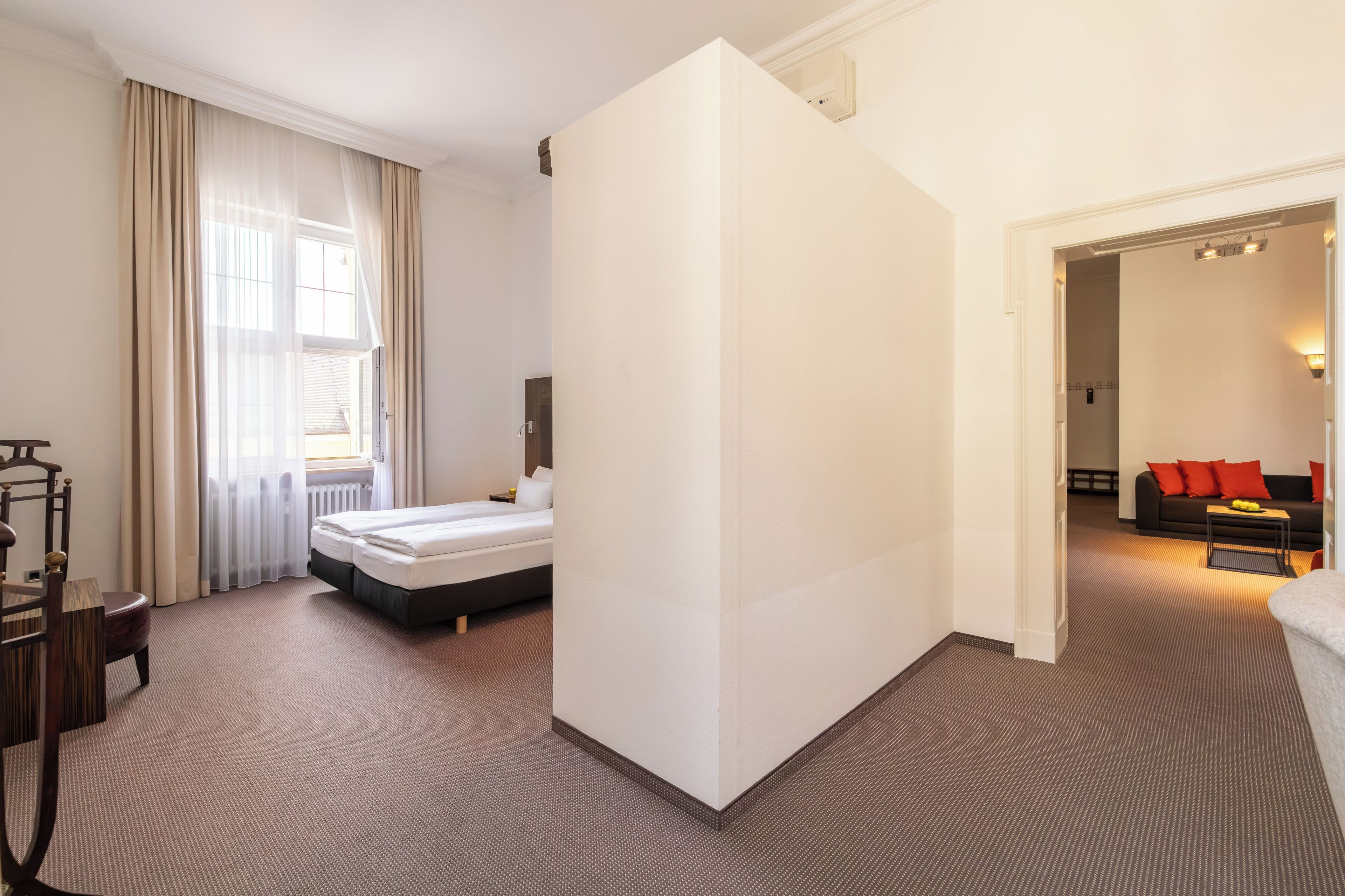 Quarto de hotel com cama, escrivaninha, cadeira, janela com cortinas e vista para outro quarto com sofá e mesa.