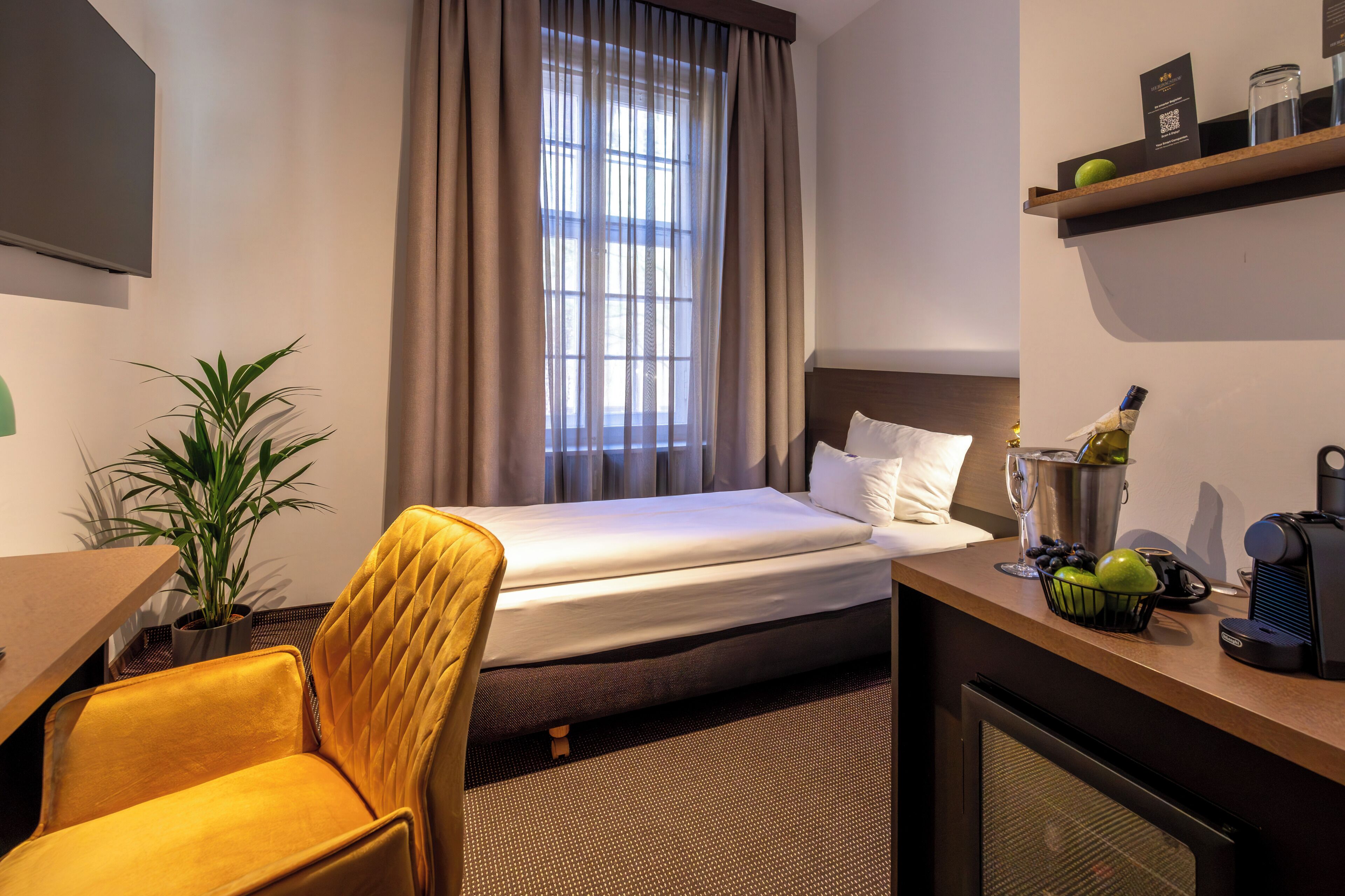 Habitación de hotel con cama, escritorio, silla, televisor, ventana con cortinas, planta y minibar.