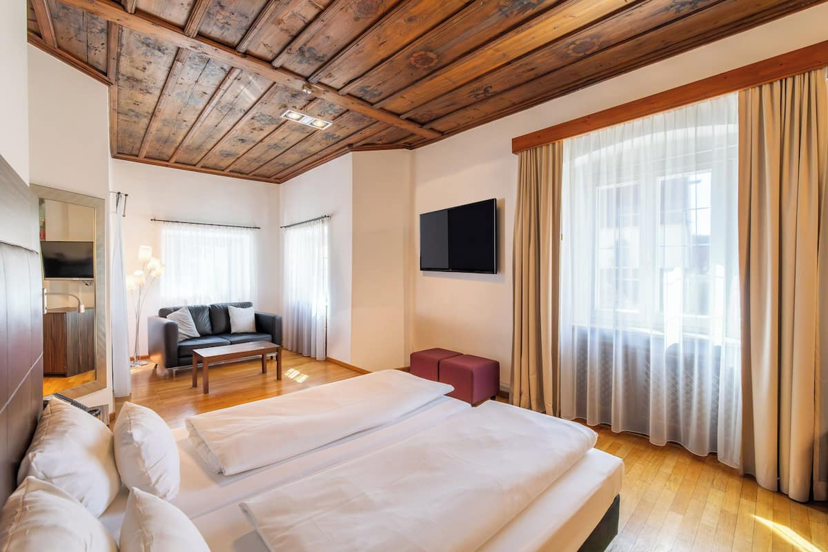 A hotel room with a wooden ceiling, a bed with white linens, a sofa, a TV, and a window with curtains.