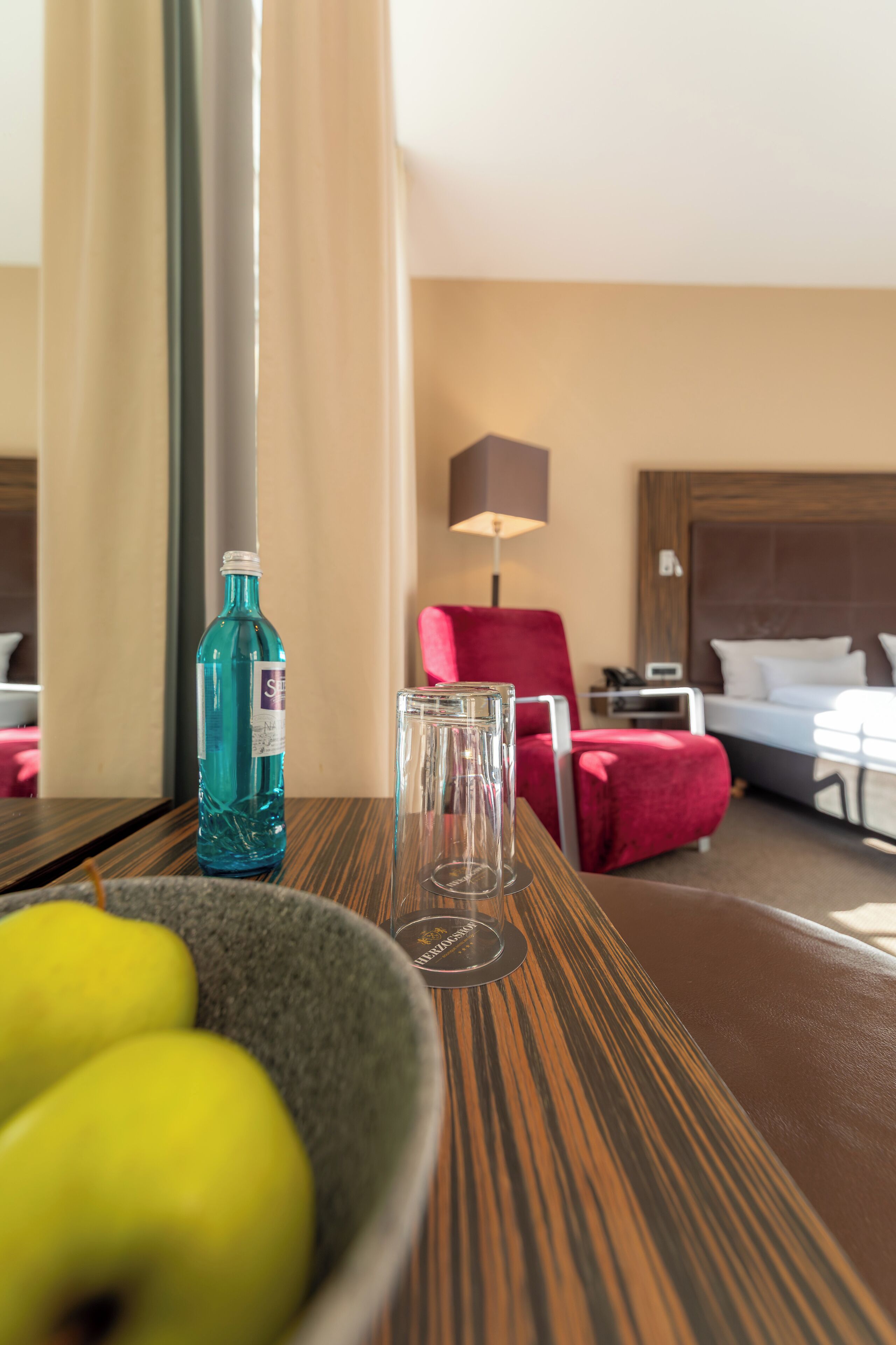 Habitación de hotel con una cama, un sillón rojo, un vaso de agua y una botella de agua sobre una mesa de madera.