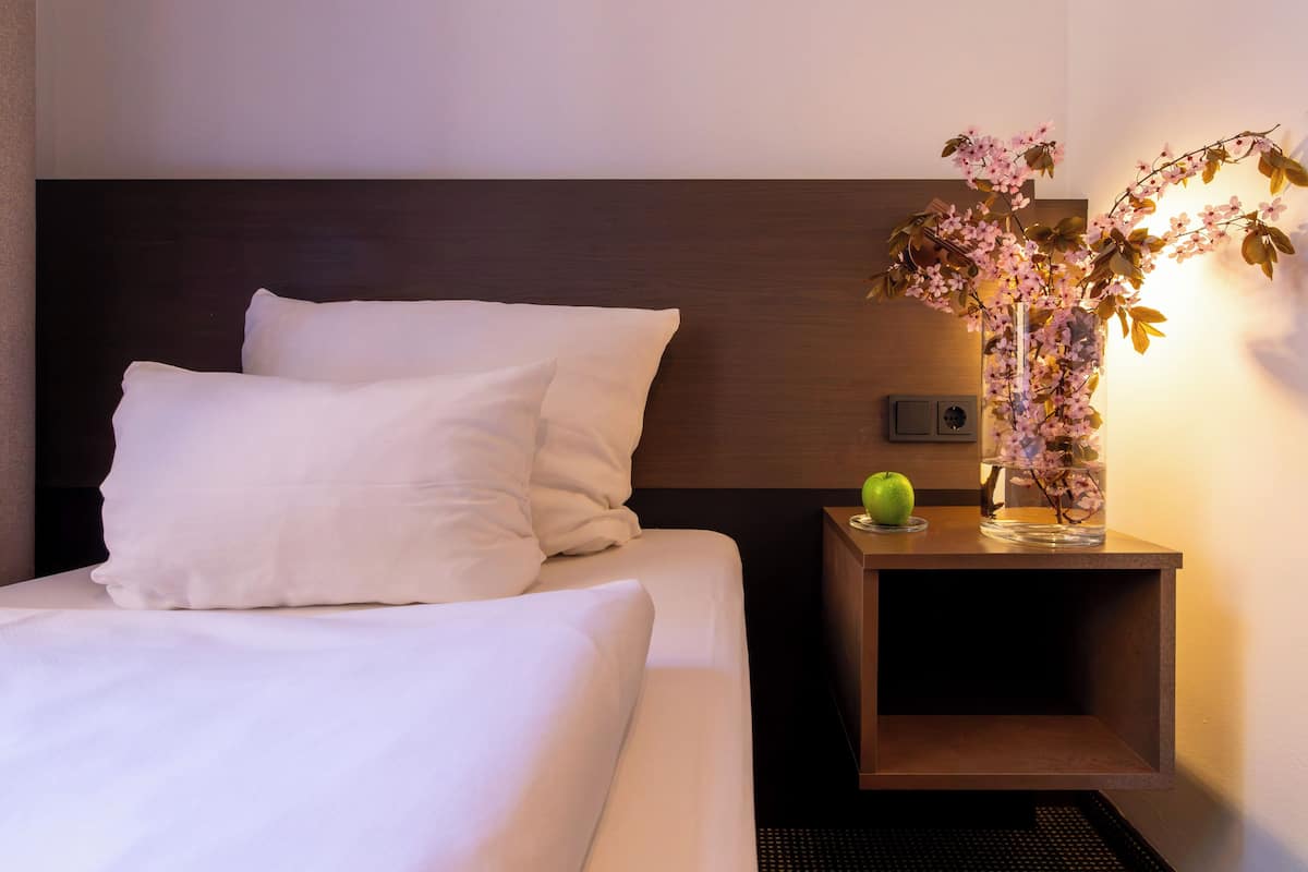 A neatly made bed with white linens, a wooden headboard, a bedside table with a vase of flowers, and a green apple on the table.