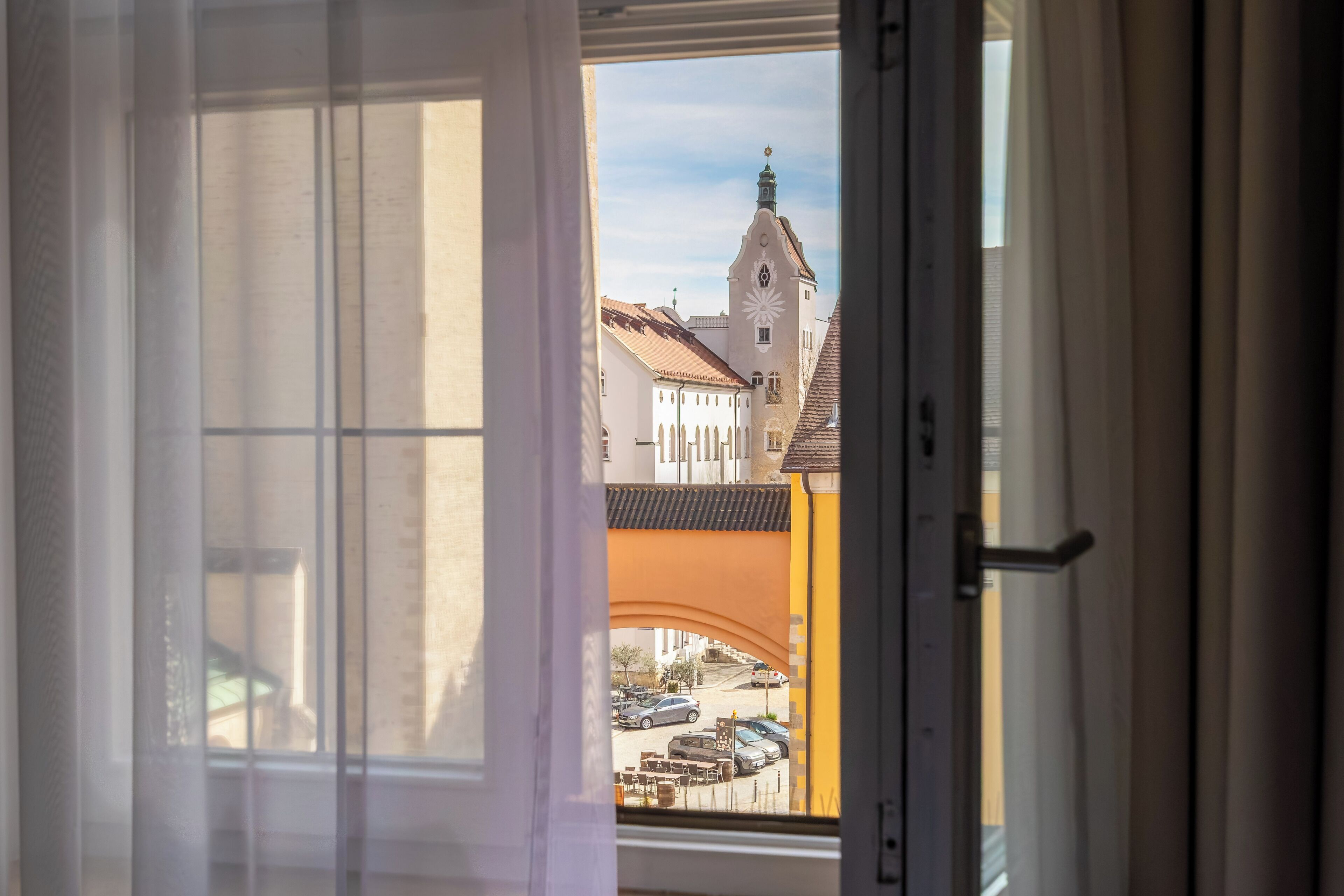 A view through an open window of a building with a clock tower, a yellow arch, and parked cars.
