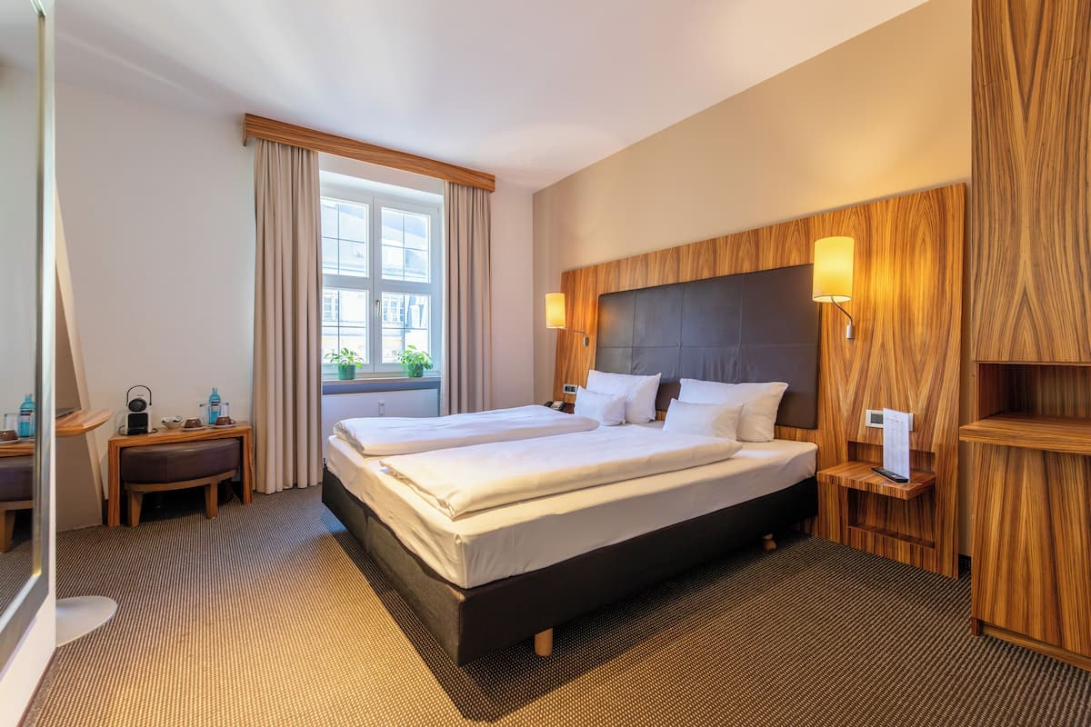 A hotel room with two beds, a wooden headboard, a bedside table with a lamp, a small desk with a chair, and a window with curtains.