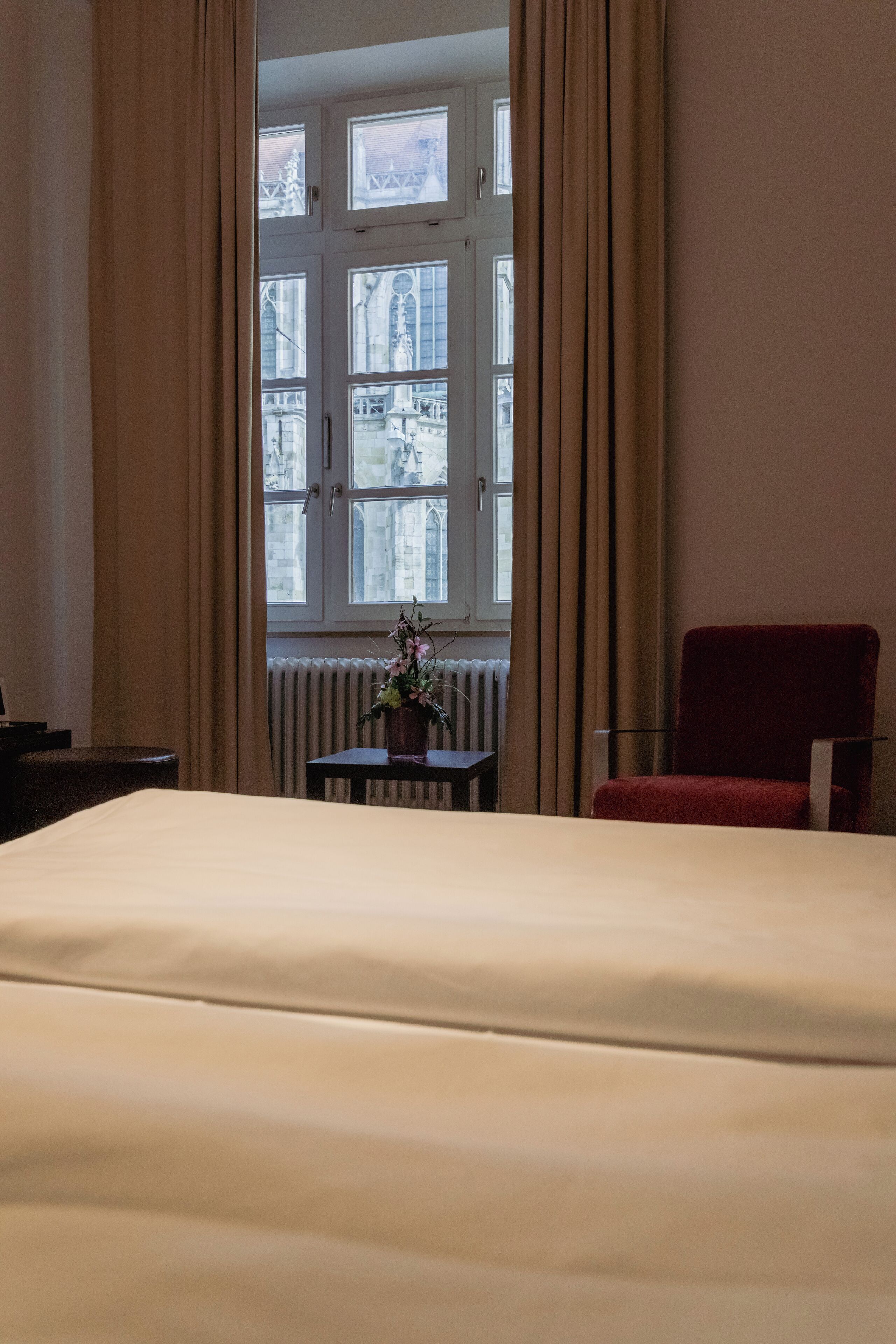Une chambre d’hôtel avec un lit, une chaise rouge, une petite table avec un vase et une fenêtre avec des rideaux.
