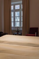 A hotel room with a bed, a red chair, a small table with a vase, and a window with curtains.