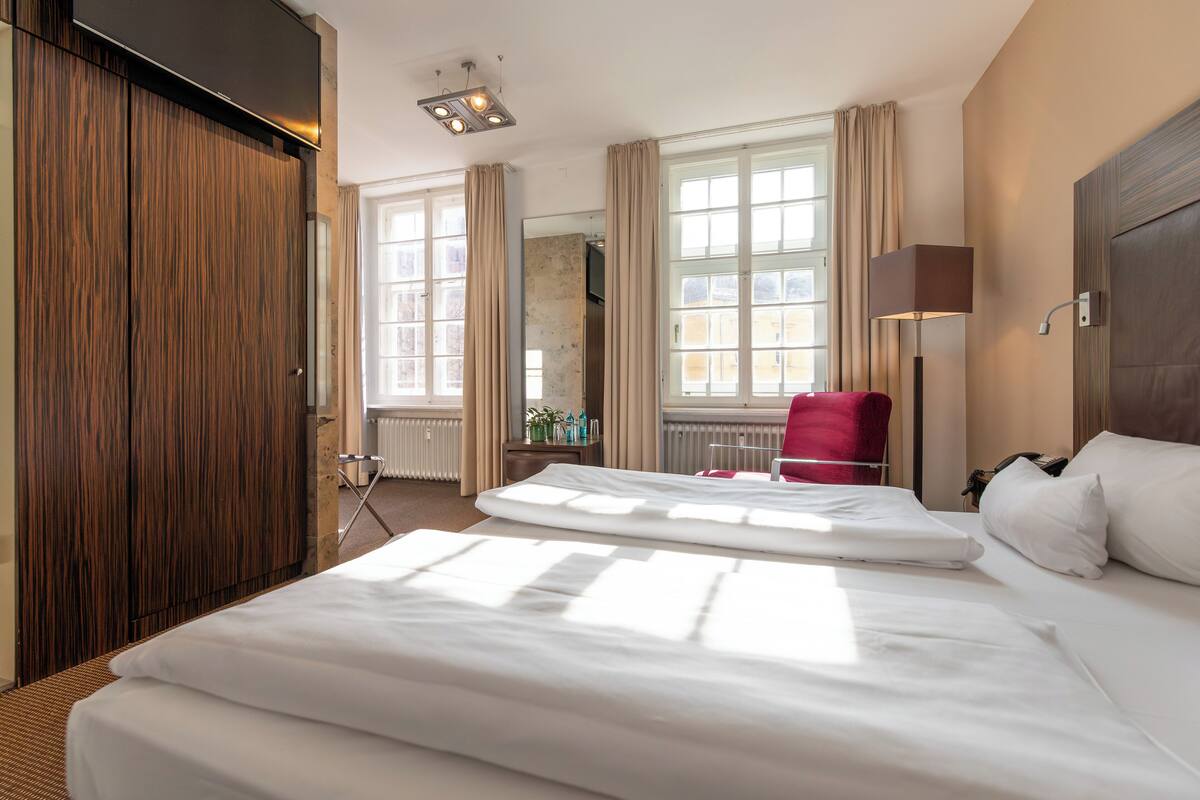 A hotel room with two beds, a red chair, a lamp, and large windows with curtains.