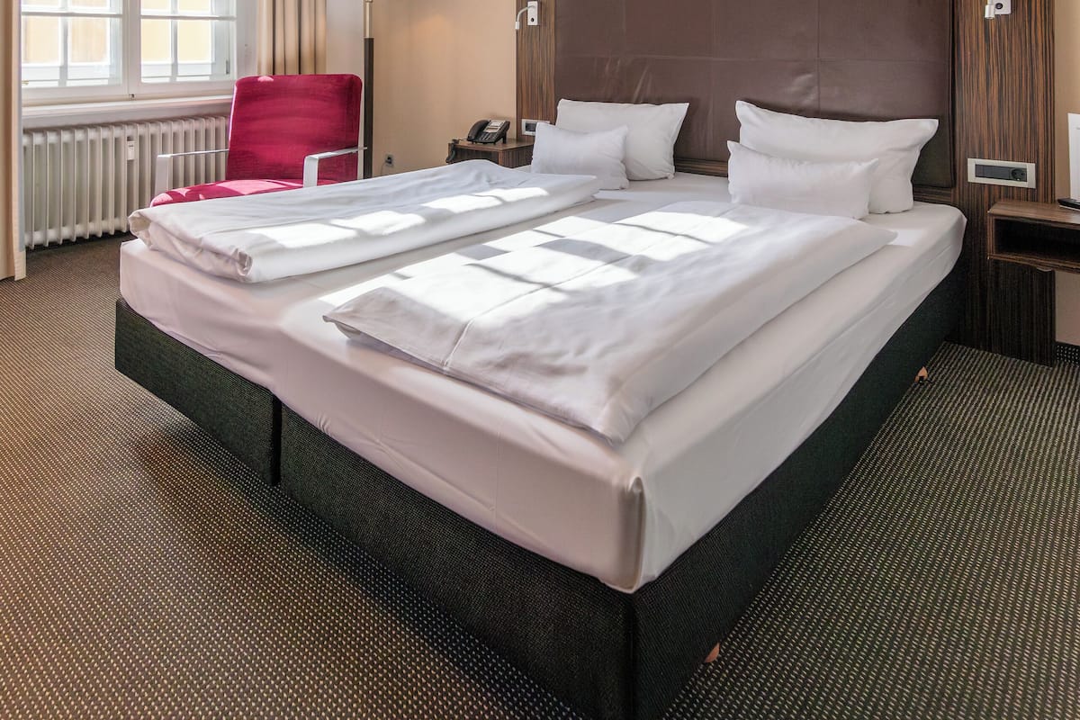 A hotel room with a large bed, a red chair, a bedside table with a lamp, and a window with curtains.