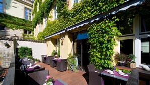 An outdoor dining area with wicker furniture and purple tablecloths, surrounded by greenery and a building with ivy-covered walls. - Hotel Regensburg Herzogshof am Dom (Regensburg)