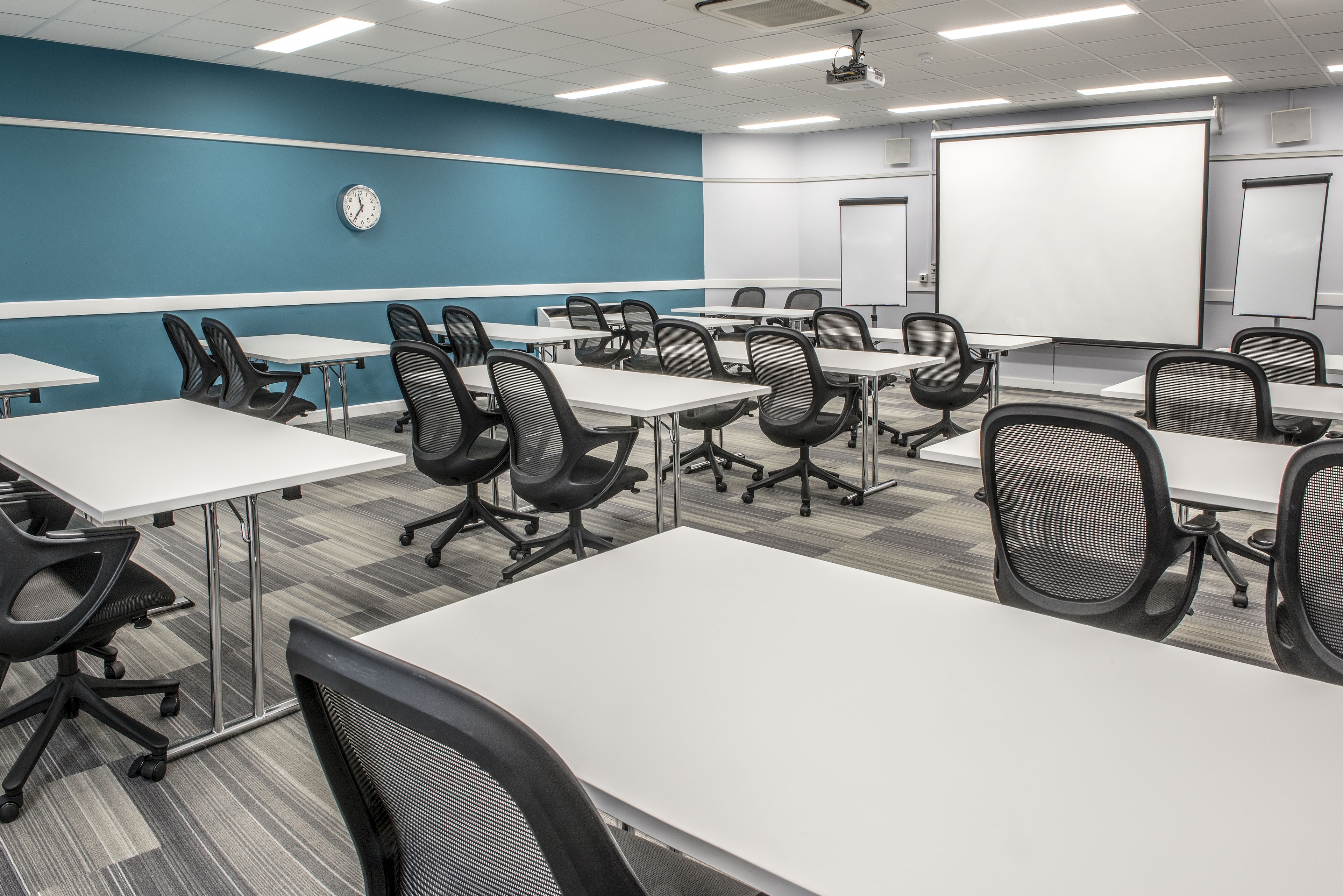 A conference room with white tables and black chairs, a clock on the wall, and a projector screen.