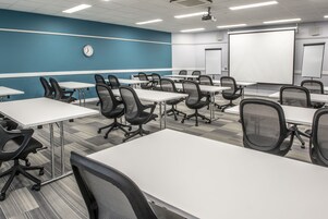 Un salón de conferencias con mesas blancas y sillas negras, un reloj en la pared y una pantalla para proyector.