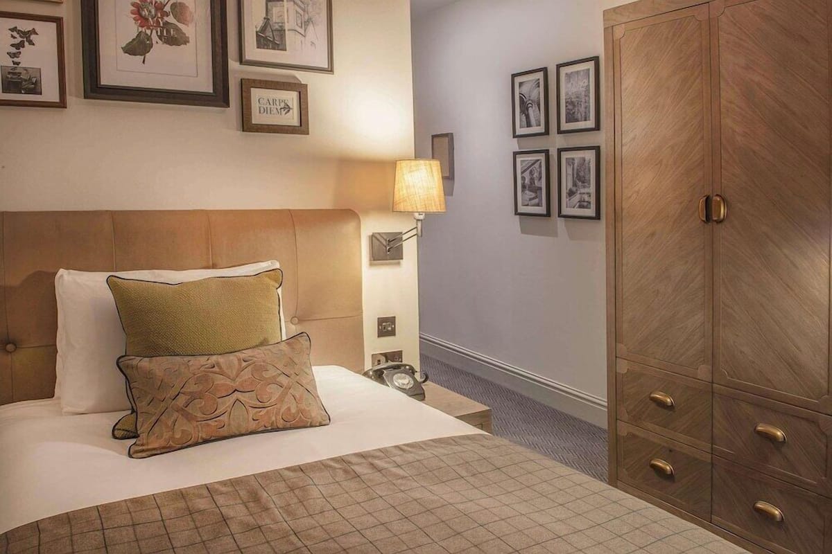 A hotel room with a bed, a nightstand, a lamp, a wardrobe, and framed pictures on the wall.